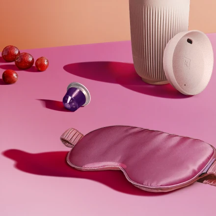 A composition of pink sleep mask with grapes, coffee capsule, and reusable cup on a pink surface.